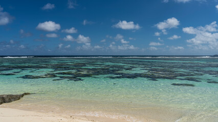 Corals are visible through the clear turquoise water of the ocean. The foam of waves over the reef in the distance.  White sand of the beach. Clouds in the blue sky. Seychelles. La Digue.
