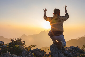 Young male kneeling down with holding christian cross for worshipping God on top rock mountain at...