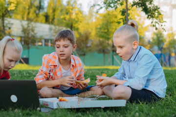 cute caucasian children sitting on grass in park having lunch eating pizza outdoors after school.