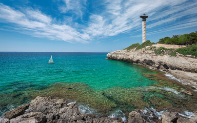 Fototapeta na wymiar Lighthouse, Sailboat and Cliffs in Torredembarra on a Shore of The Mediterranean Sea. Tarragona, Catalonia, Spain.