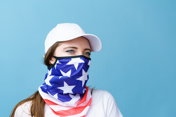 Young woman with America flag mask on her face in white t-shirt and cap for mockup looking at camera on blue studio background