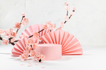 Romantic spring scene with pink cylinder podium mockup, branch lush pink sakura flowers in sunlight, japan fans in light white interior, template for presentation cosmetic products, goods, branding.