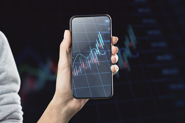 Close up of female hand holding mobile phone with glowing forex chart on blurry dark grid background. Trade, finance and invest concept.