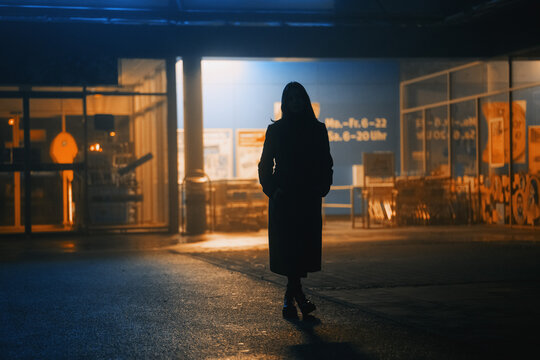 dark silhouette of a girl dressed in a long coat against the background of a night city with advertising showcases