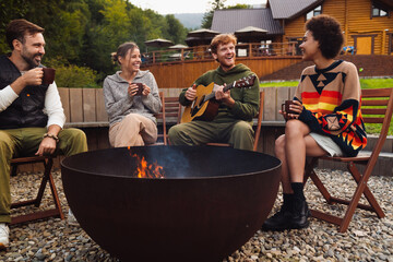 Young friends drinking tea and playing guitar while sitting around campfire in nature