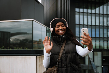 Fototapeta premium Positive afro woman making video call via mobile phone while standing outdoors near modern office building