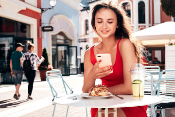 Young beautiful smiling hipster woman in summer red dress. Positive model sitting in veranda cafe in street. Female drinking fresh cocktail smoothie drink in plastic cup with straw. Eats sweet cake