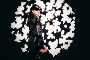 Obraz premium Young beautiful brunette female in trendy evening reflecting dress. Sexy carefree woman posing near white wall in studio in circle a of shadows from tree light. Fashionable model in sunglasses