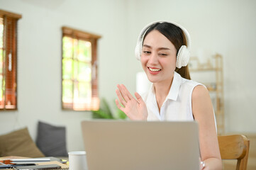 Beautiful Asian businesswoman wearing headphones, waving hand, talking, having a video call