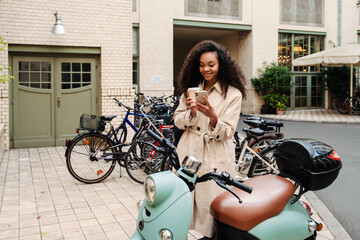 Fototapeta na wymiar Smiling african girl drinking coffee and using smartphone while standing outdoors at bike parking