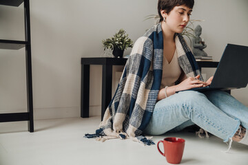 Young woman wrapped in plaid working on laptop while sitting on floor