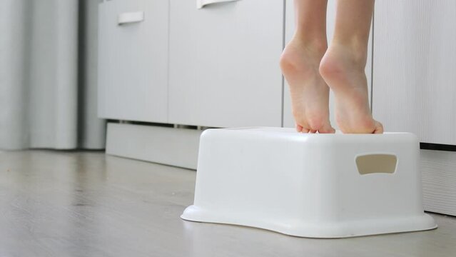 Small Children's Legs Getting Up On The Step. Small Children's Legs Stand On A Special Children's Stand On Kitchen. Toddler Girl's Feet Standing On Step Stool In Kitchen. Small Child Helps At Home