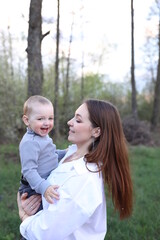 Fototapeta premium mother hugs her little son in spring near a blooming tree. Spring and plum blossom. Mother's Day. The love of a son and a mother. Gray clothes for a one-year-old boy. Kiss the son. child's laughter
