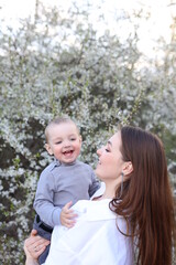 mother hugs her little son in spring near a blooming tree. Spring and plum blossom. Mother's Day. The love of a son and a mother. Gray clothes for a one-year-old boy. Kiss the son. child's laughter