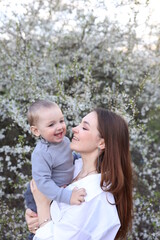 mother hugs her little son in spring near a blooming tree. Spring and plum blossom. Mother's Day. The love of a son and a mother. Gray clothes for a one-year-old boy. Kiss the son. child's laughter