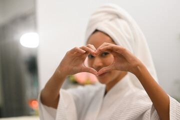 Youthful asian woman doing heart symbol shape with hands smiling to her reflection in mirror. Beauty and self care concept.