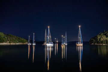  white boats located over horizon, colorful lights coming from yachts reflect on the surface of the the Gulf sea. Shot at blue hour in Agios Stefanos Harbour, Corfu , Greece