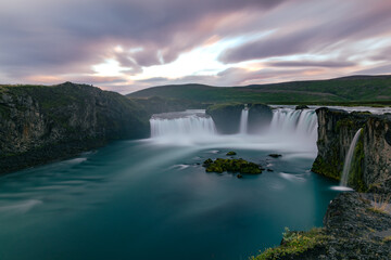 Fototapeta premium Long exposure sunset landscape over the icelandic Godafoss waterfall with blurred water falling into turquoise river and cloudy majestic sky