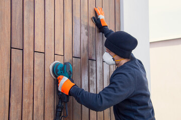 Man in dust mask sanding teak cladding on the house facade, exterior wood siding renovation