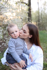 mother hugs her little son in spring near a blooming tree. Spring and plum blossom. Mother's Day. The love of a son and a mother. Gray clothes for a one-year-old boy. Kiss the son. child's laughter