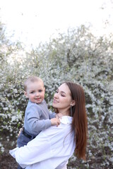 Obraz premium mother hugs her little son in spring near a blooming tree. Spring and plum blossom. Mother's Day. The love of a son and a mother. Gray clothes for a one-year-old boy. Kiss the son. child's laughter