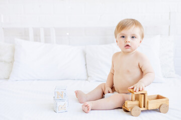 cute blonde baby boy is sitting on a white bed at home in diapers and playing with a wooden toy car