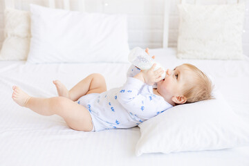 cute baby with a bottle of milk sucks holding it in his hands lying on her back on a baby bed in a children's bright room, baby food concept