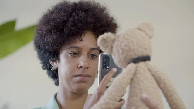Young lady taking picture of teddy bear toy using her smartphone. Closeup shot of pretty Black woman with short curly hair holding soft toy and looking at screen. Photography, hobby, startup concept