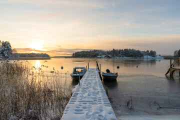 Fototapeta premium Soukka, Finland - 09.12.22: Winter sunset over the snow-covered islands in the bay. Frozen bay. Pier with boats in the foreground. Winter landscape. Scandinavia.
