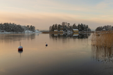 Naklejka premium Soukka, Finland - 09.12.22: View from the shore of a small snow-covered island with houses. Frozen bay. Winter landscape. Scandinavia.