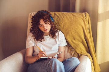 Curly-haired young girl in headphones sitting in the armchair and making notes in a diary