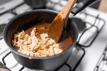 A young woman is mixing chicken with mole sauce using a gas stove