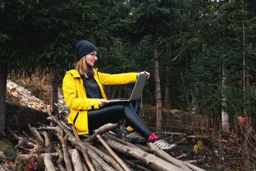 Woman wearing glasses and yellow jacket working on laptop outdoors surrounded by beautiful nature, forest and mountains. Workation concept. Freelancer work, remote work