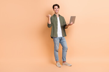 Full body length photo of young man interested looking empty space pointing finger with netbook promotion ad isolated on beige color background