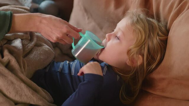 Close up, woman hand gives warm drink to her child on the sofa with fever, mothers helping hand, caring tender family moment. Sick kid, corona symptoms, virus disease, ilness concept. HQ 4K footage
