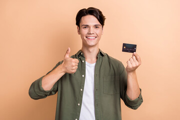 Photo of positive person hold plastic debit card demonstrate thumb up isolated on beige color background