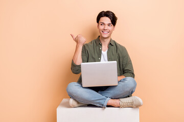 Full body photo cadre of young optimistic it developer guy sitting with laptop look point finger mockup isolated on beige color background
