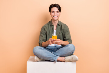 Full body length photo of young student sitting platform cube use his smartphone chatting online with friends isolated on beige color background