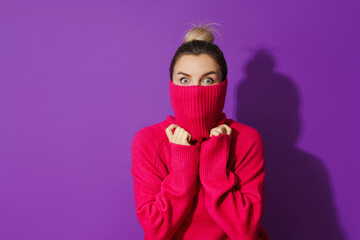 Young woman is hiding her face inside a warm and cozy polo neck sweater against purple background