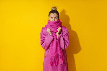 Young cheerful woman wearing pink coat and wool scarf against yellow background