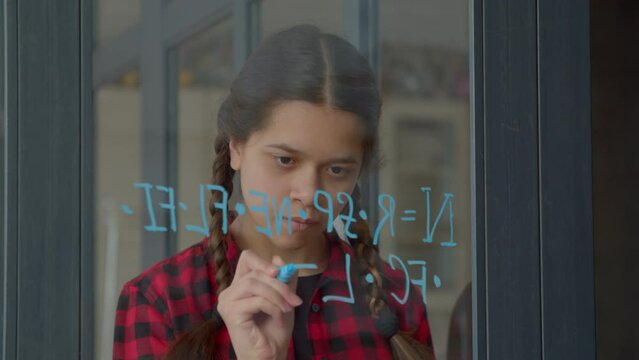 Smart cute Latin teenage girl with pigtails writing with felt tip pen equation on window glass, estimating number of active, communicative extraterrestrial civilizations indoors.