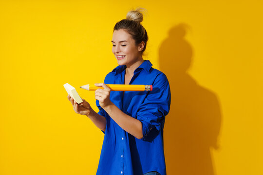 Cheerful woman wearing blue shirt holding giant pencil on yellow background