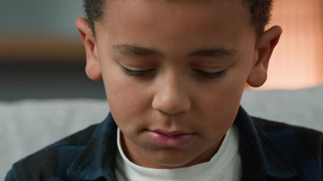 Close up sad upset little African American boy child looking down with guilty face ashamed. Headshot disappointed ethnic kid alone schoolchild schoolboy baby with closed eyes dreaming praying dream