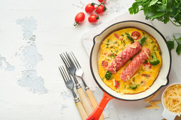 Omelette or frittata with two sausages, melted cheese, cherry tomatoes, broccoli and parsley on red skillet or iron pan on white old concrete background. Healthy breakfast food concept. Top view.