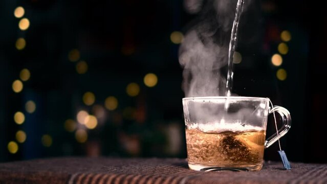 Pouring hot water in a transparent glass mug. Making seasonal drink with triangular tea bag. Cozy Christmas mood with steaming rooibos tea in backlit. Evening lights, bokeh background and copy space.