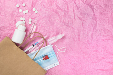 Medical items such as ampoule pills, a syringe and a mask peek out of a paper bag against a background of pink crumpled paper.  Lots of empty space for text