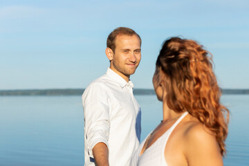 Happy smiling Caucasian man holds hand of Latin woman, blue sky and water on background. International couple in love look at each other. Romantic sunny day. St Valentine's day,enjoying togetherness