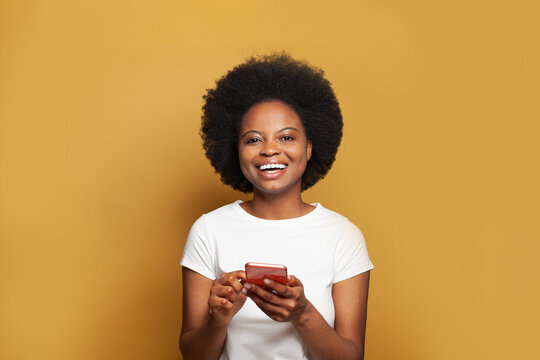 Surprised happy brunette woman in white t-shirt using smartphone while looking at the camera with friendly smile over yellow wall background