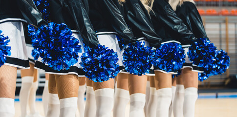 Blue pom-poms being held by the cheerleaders standing in line. Sports hall blurred in the background. High quality photo