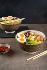 Two bowls of ramen soup with chicken breast, vegetables, mushrooms, egg and chopsticks on a gray concrete background. Front top view, copy space, healthy eating concept..